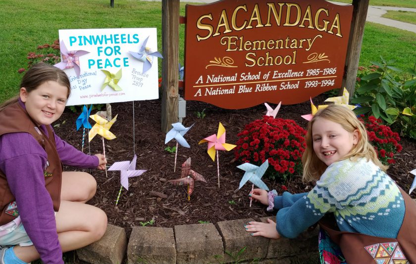 two students kneeling in front of the school sign with the pinwheels they made in the ground