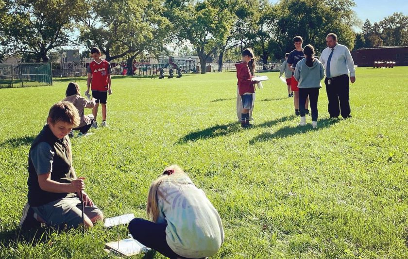 students on field of grass with teacher. some are sitting and others are standing in a group