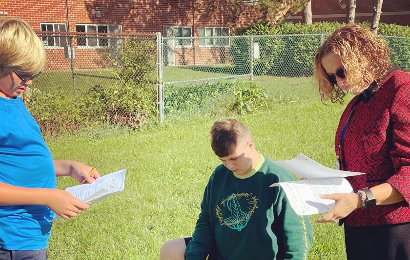 a student kneeling on grass another student is standing looking at a paper and an adult has papers in her hand