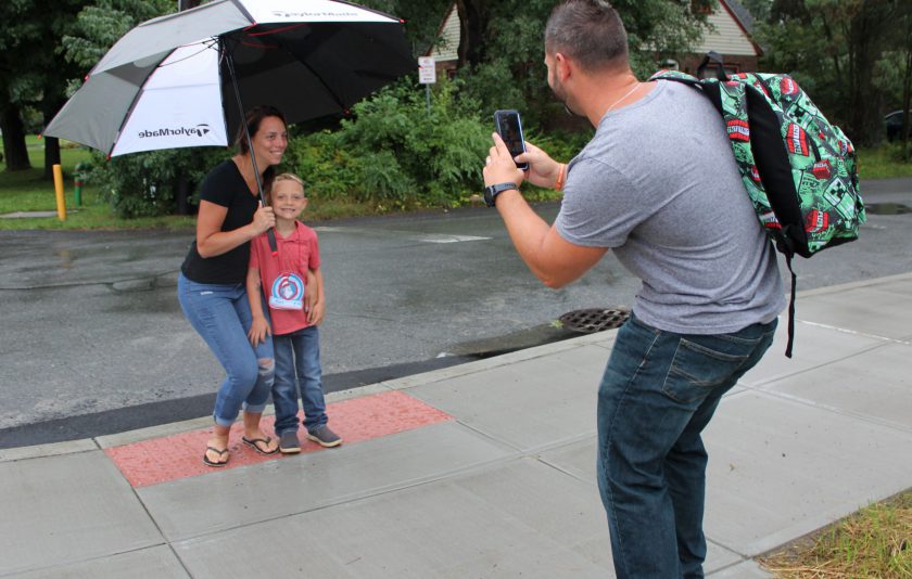 woman and child stand under umbrella while man wearing backpack takes a phone photo