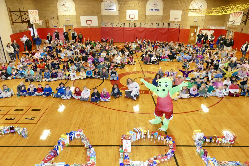 students sitting all around the gym floor with dragon mascot jumping and food collected spelling out care