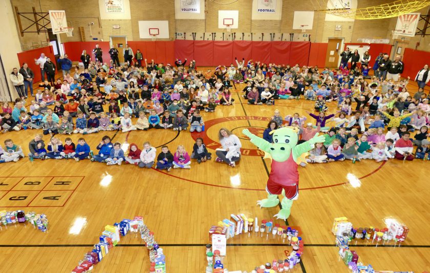 students sitting all around the gym floor with dragon mascot jumping and food collected spelling out care