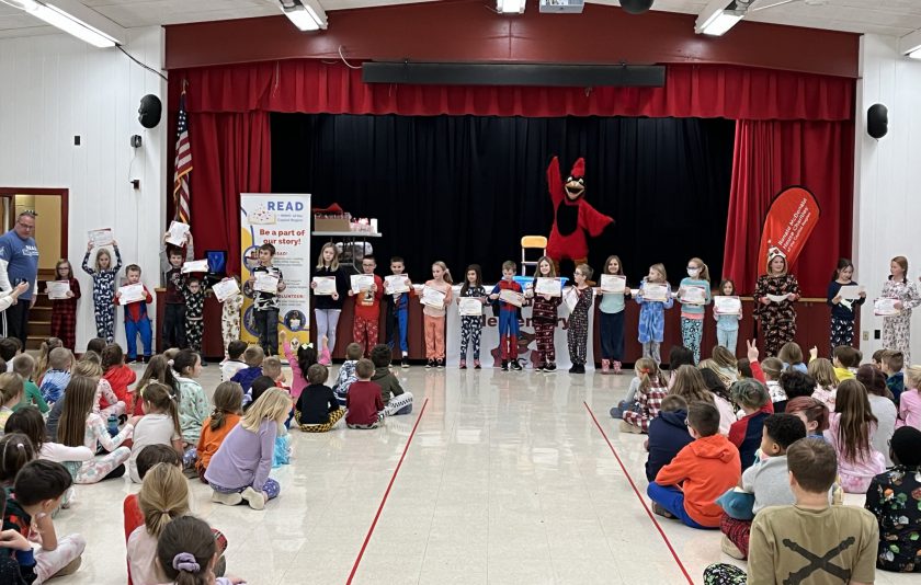 students stand by stage holding certificates while other students are seated in front of them