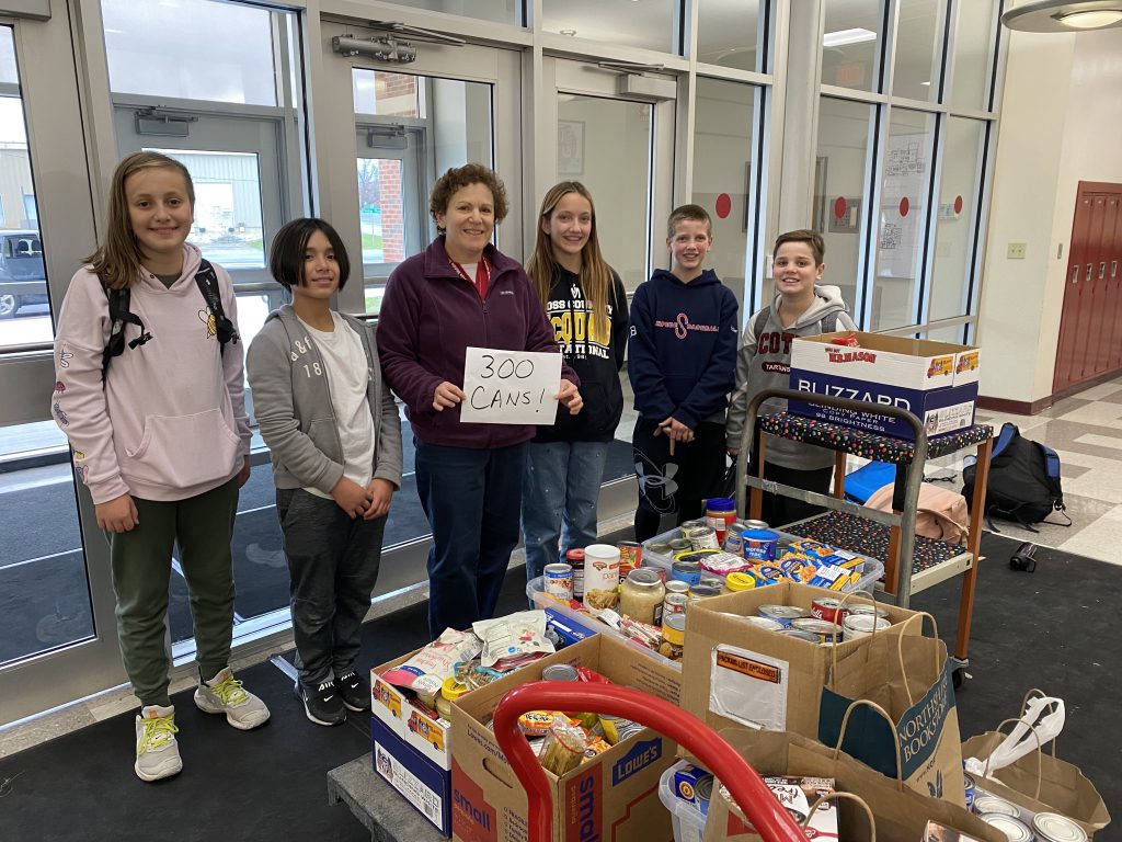 six students standing in front of boxes of food items in cans. A teacher is holding a sign that says 300 cans!