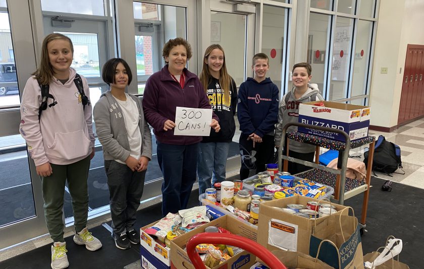 six students standing in front of boxes of food items in cans. A teacher is holding a sign that says 300 cans!
