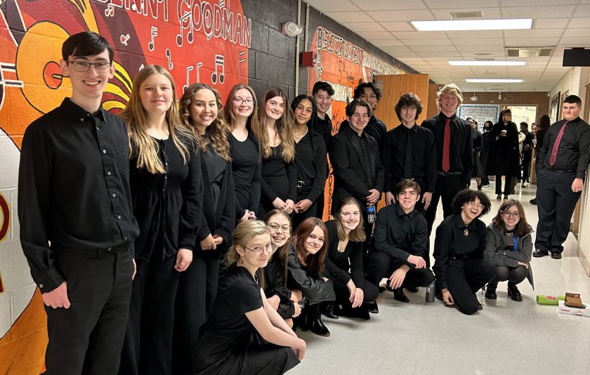 18 music students dressed in black and standing in front of a colorful wall in a hallway. It's a design using yellows and reds.