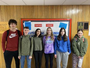 Six students stand in a line smiling. the first student is significantly taller than the other five who are about the same height. 