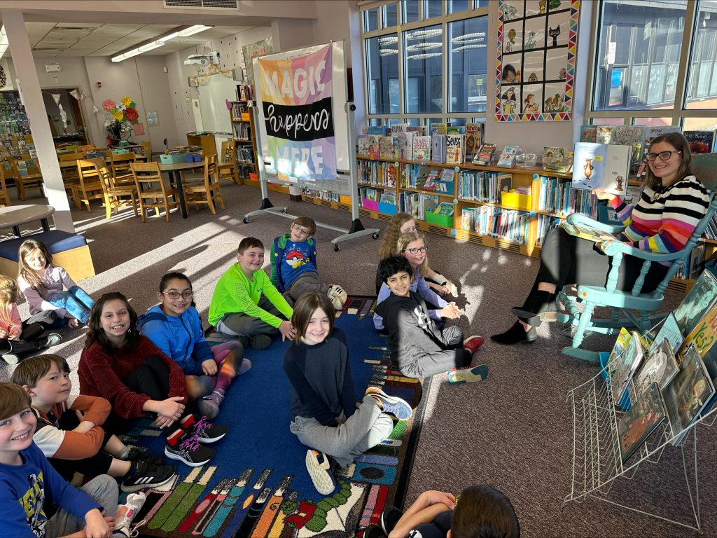 teacher sitting on a chair holding a book in the library and students are sitting on the floor in front of her