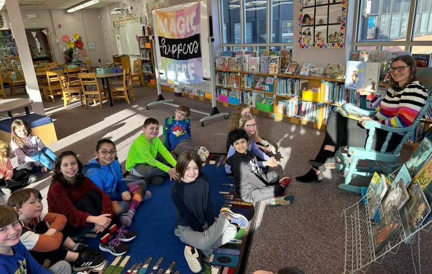 teacher sitting on a chair holding a book in the library and students are sitting on the floor in front of her