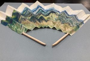 a thick material folded like a fan with sticks on both sides of the material. the pattern starts at the top with white then shades of blue, and greens and browns and yellows.