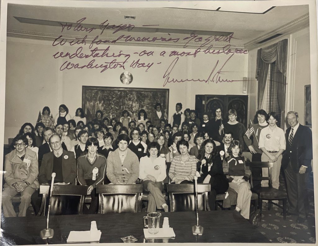 an old photo from 1981, where students and adults are sitting together in chairs and some are standing looking forward at the camera