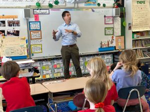 man in blue shirt standing in front of a whiteboard talking to students who are sitting in their desks