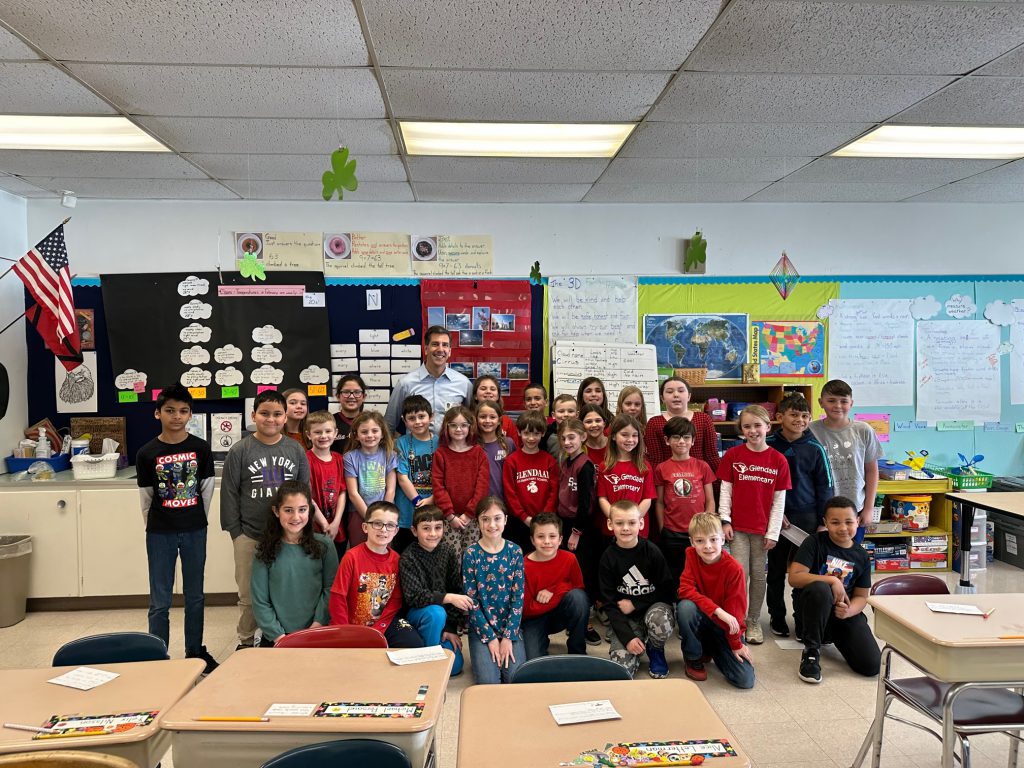 a classroom with a hanging American flag and a bunch of students gathered around a man in a light blue shirt - weatherman Steve LaPointe