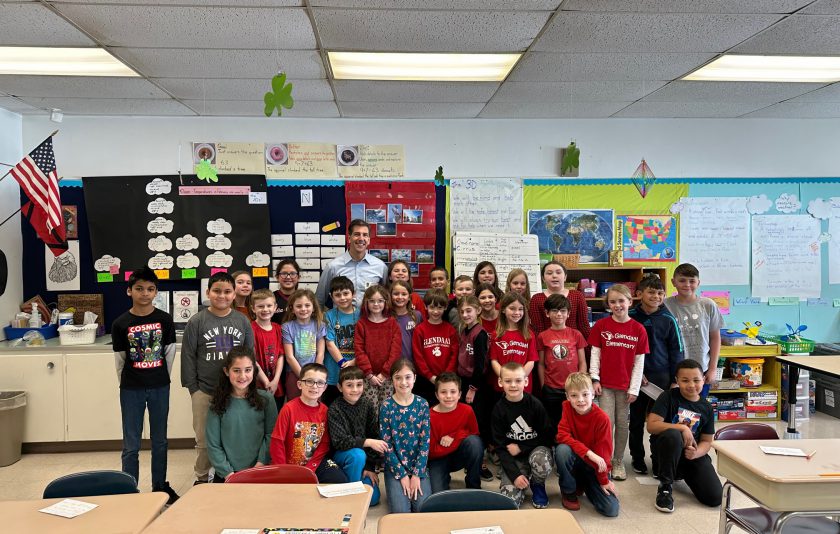 a classroom with a hanging American flag and a bunch of students gathered around a man in a light blue shirt - weatherman Steve LaPointe