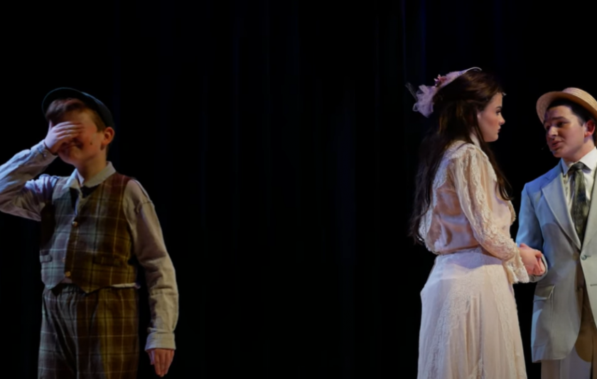 three people on a stage with a black background, a woman in a white dress holds hands with a man in a suit and top hat, and a child has his back toward them with his hand over his eyes