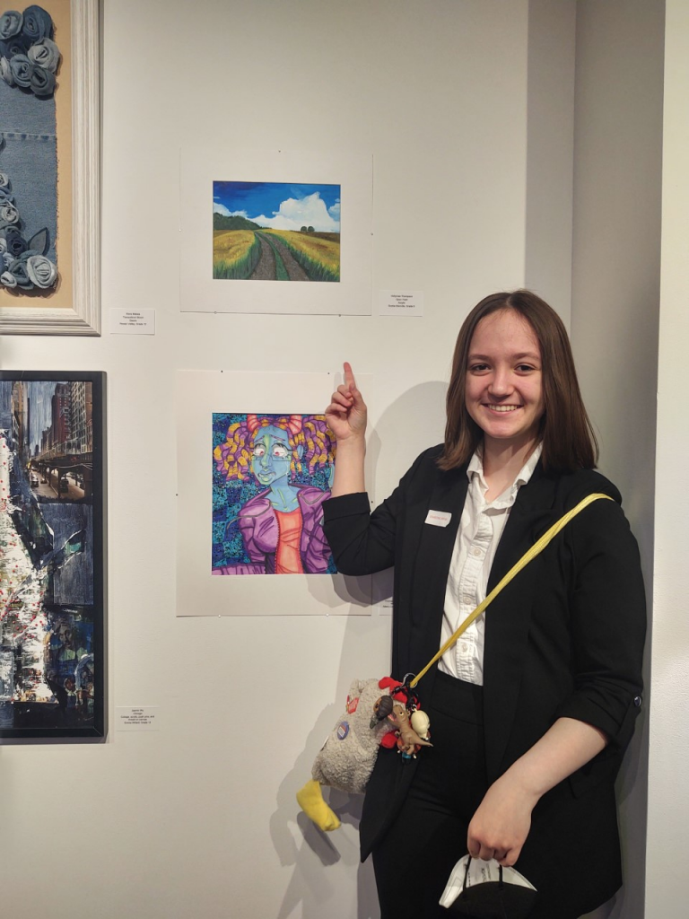 students stands near her artwork and points up at it on the wall