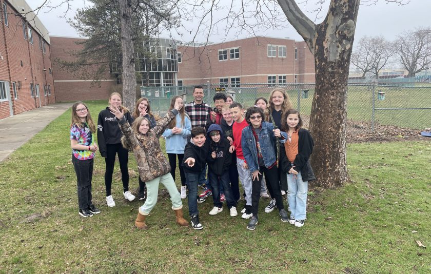 15 students standing around their teacher outside in the schoolyard