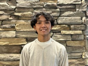 studenr with dark wavy hair and a beige shirt is smiling in front of a stoned wall