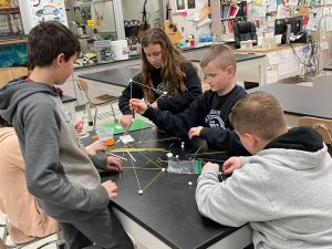 four students standing around a table designing a shape with sticks 