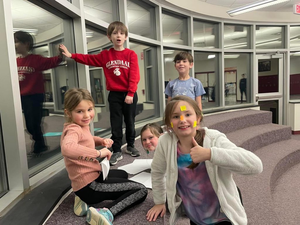 five students standing and sitting on steps near a window, one student has yellow sticky notes on her face and she is giving a thumbs up