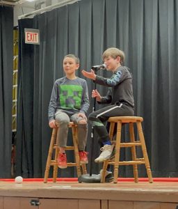 Two students sitting on stools in front of a microphone entertaining a crowd