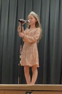 students standing on stage with a mic in her had is wearing a sequin dress and a crown