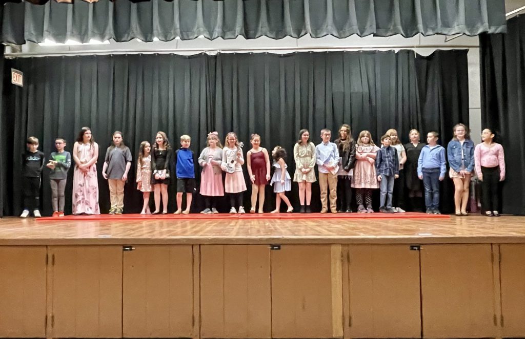 21 students standing on stage in a line looking out to the crowd