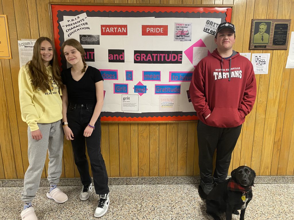 two students standing next to each other turned inward, another students is standing further away behind a black dog