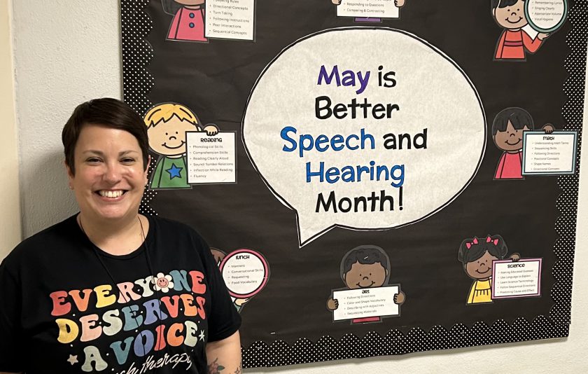 adult wearing a shirt that say everyone deserves a voice as she stands near a bulletin board that says May is better speech and hearing month