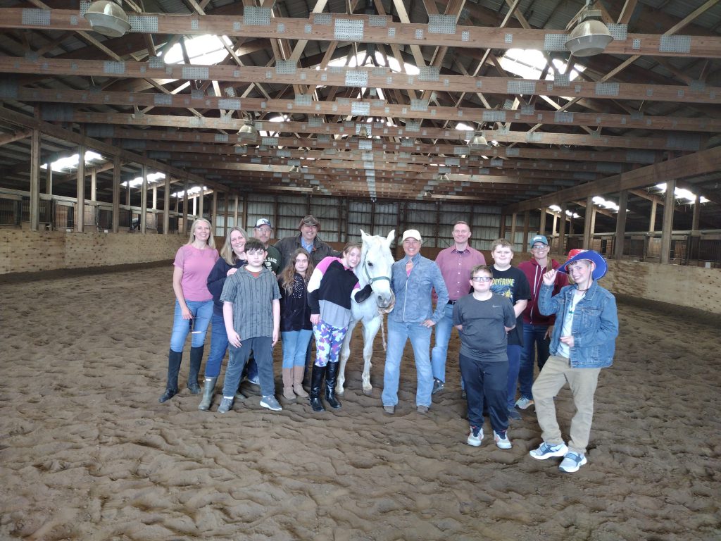 in a barn, a white horse is surrounded by smiling students and adults
