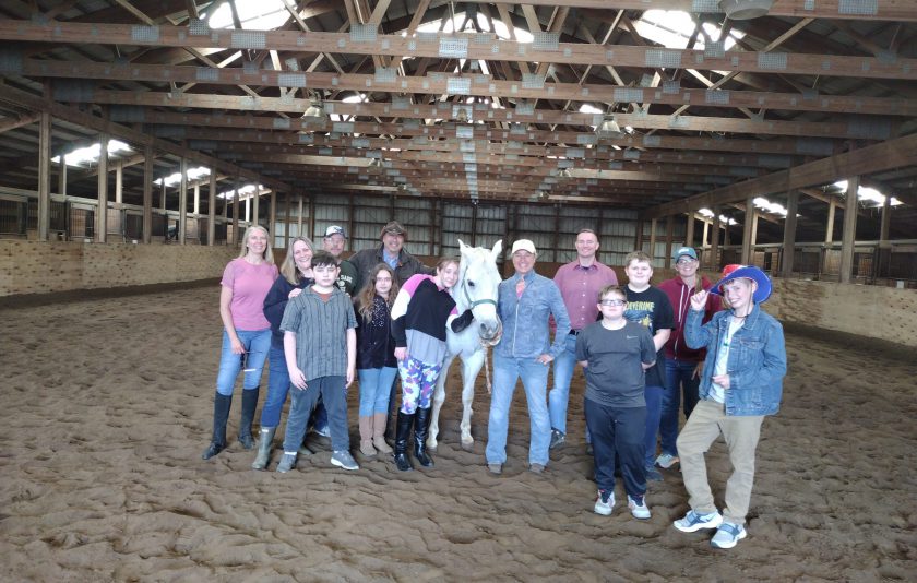 in a barn, a white horse is surrounded by smiling students and adults
