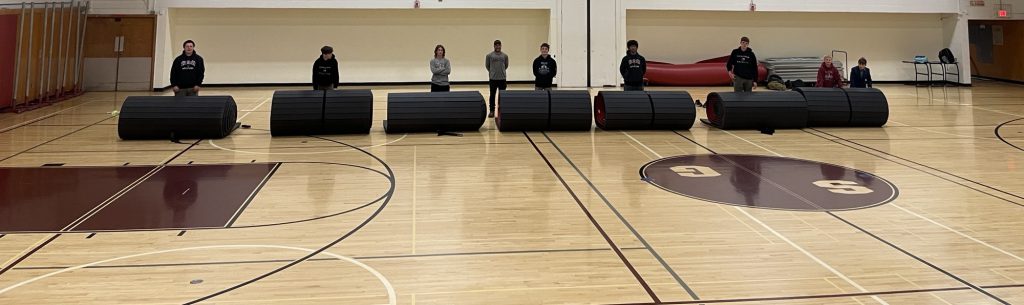 students standing behind rolled up mats on a gym floor