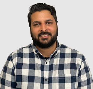 Man with black hair and a beard and moustache. He is wearing a black and white checkered shirt