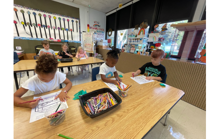 three students coloring together at a table and there are three other students coloring at a table behind them