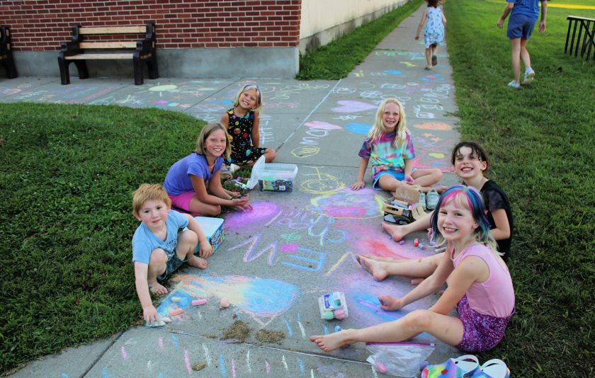 six students sitting on the ground outside school drawing with chalk