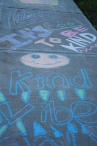 sidewalk chalk drawing in blue, orange, purple, green, pink, white, and yellow. There's a smile face and the words It's cool to be Kind 
