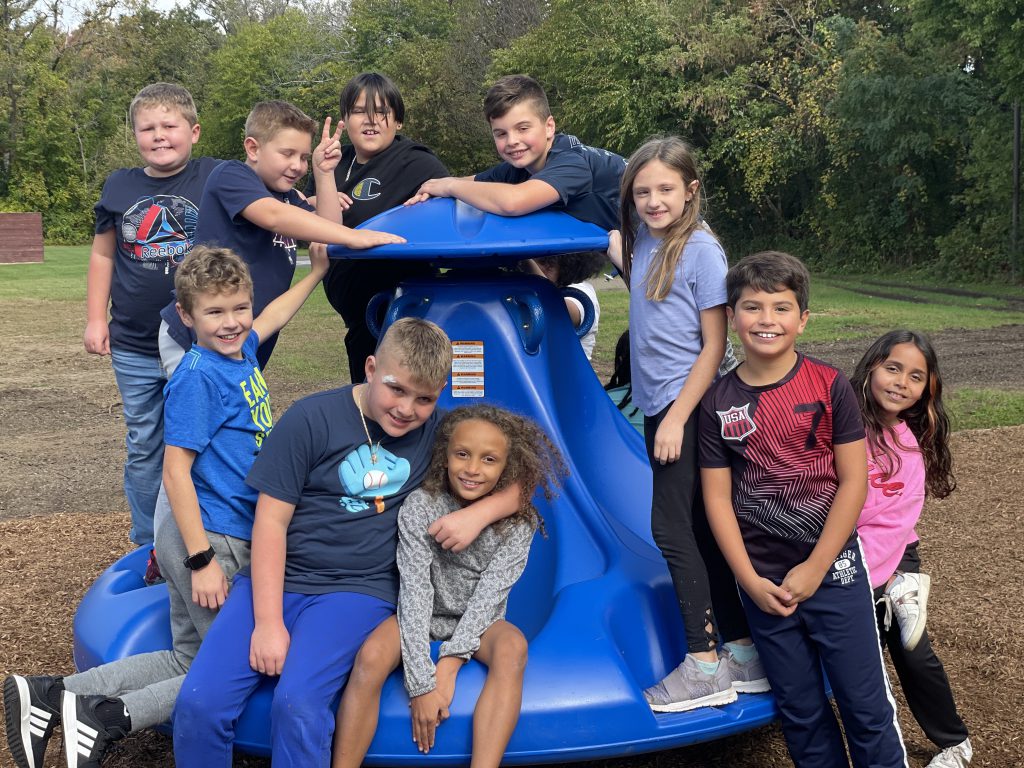 students sitting on and standing around ten spin playground equipment