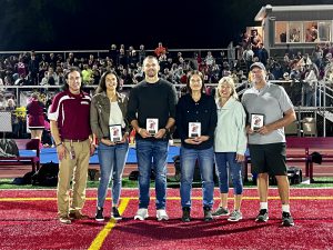 Six adults standing in a line in the football field with a crowd in the stands behind them