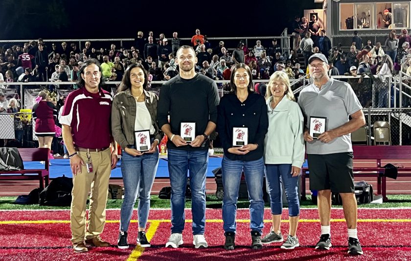 Six adults standing in a line in the football field with a crowd in the stands behind them