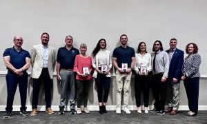 11 adults standing together. All are smiling and some are holding their awards.