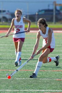 Two field hockey players in action one is watching the other player hit the orange ball with her white stick