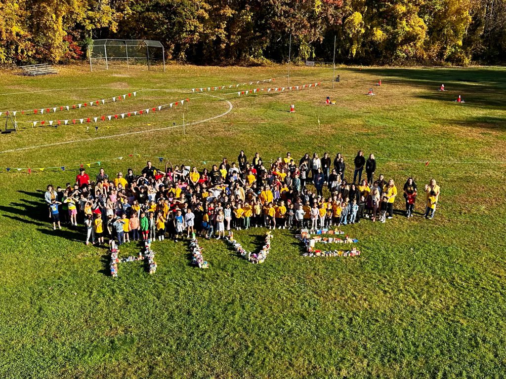 all students and faculty are outside standing on grass with food they collected making the word HIVE