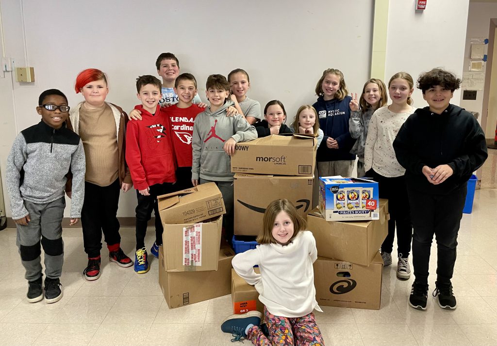 14 students standing around cardboard boxes of packed items for charity.