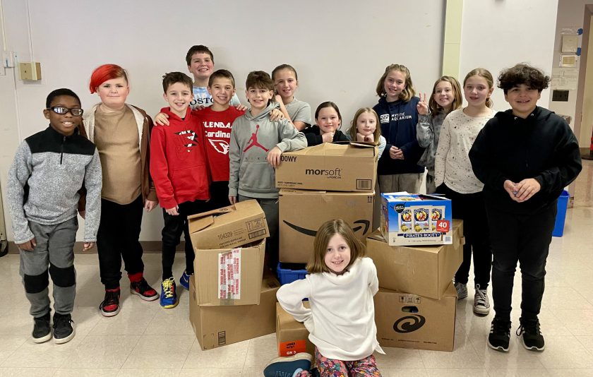 14 students standing around cardboard boxes of packed items for charity.