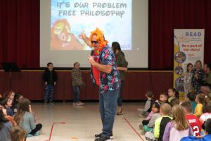 Principal wearing a colorful shirt, sunglasses, and orange lion's mane singinging for students