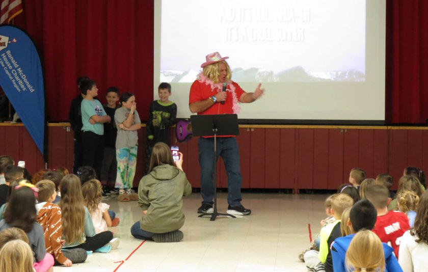 Principal dressed in a blonde wig with a pink cowboy hat and pink feathers singing for students
