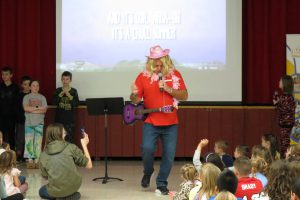 Principal dressed in a blonde wig with a pink cowboy hat and pink feathers singing for students.