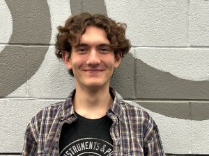 Student with short brown wavy hair wearing a plaid shirt smiling.