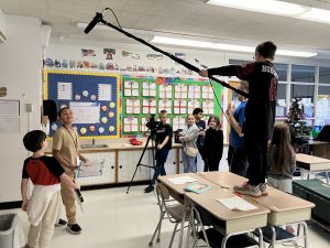 students in classroom making a movie, one is holding a boom mic, others are behind the camera or actors 
