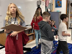 young student with long blonde hair holds clipboard and other students prepare for acting